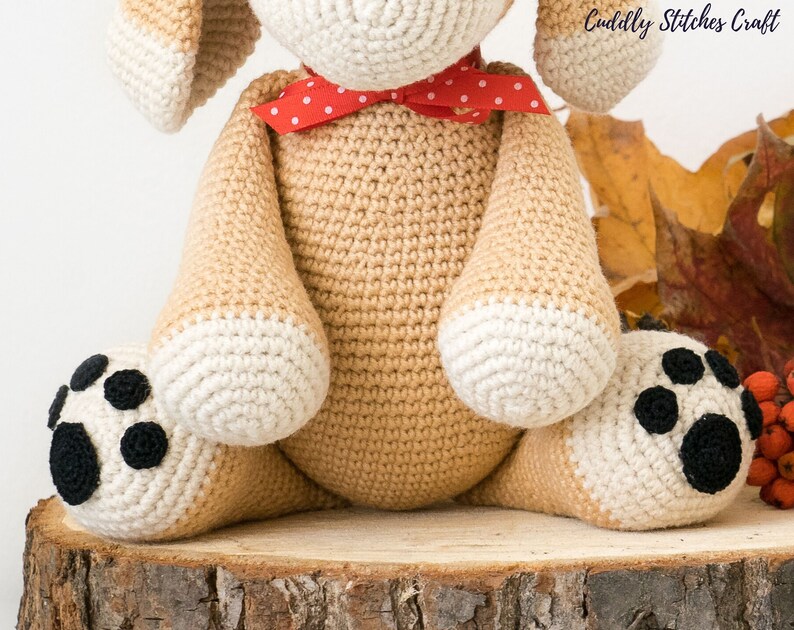 May include: A crocheted beige dog with white paws and black paw pads. The dog is wearing a red and white polka dot bow tie. The dog is sitting on a wooden log.