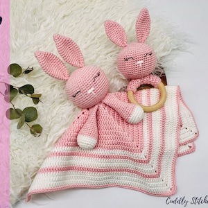 Puede incluir: Un sonajero de conejo de crochet rosa con un anillo de madera adjunto. El conejo está acostado sobre una manta de crochet rosa y blanca a rayas.