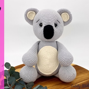 May include: A crocheted koala amigurumi toy with gray fur, a cream-colored belly patch, and black eyes. The koala is sitting on a wooden surface.
