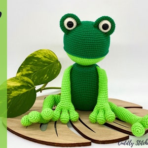 May include: Green crocheted frog amigurumi toy with black eyes, sitting on a wooden leaf-shaped base.