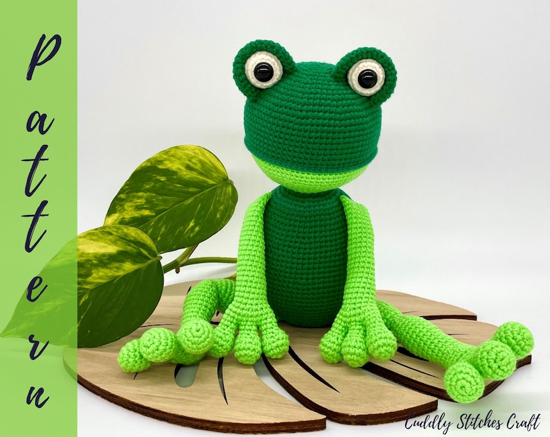 May include: Green crocheted frog amigurumi toy with black eyes, sitting on a wooden leaf-shaped base.