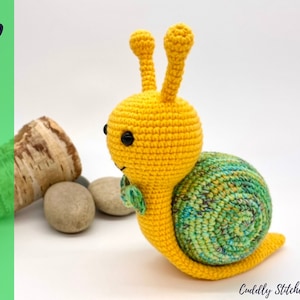Könnte beinhalten: Eine gehäkelte gelbe Schnecke mit einer grün-blauen Schale. Die Schnecke hat schwarze Augen und eine grüne Blätter-Schleife.