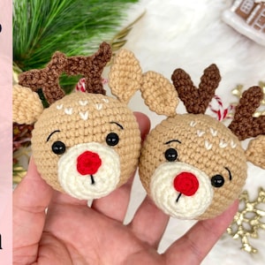 May include: Two handmade crochet reindeer ornaments. Each ornament features a tan head with a white muzzle, a red nose, and black eyes. The reindeer have brown antlers and are decorated with white stitching. The word "Pattern" is visible in the image.