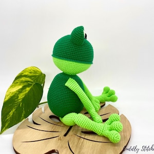 May include: A green crocheted frog sitting on a wooden leaf-shaped base. The frog has black eyes and a light green belly.