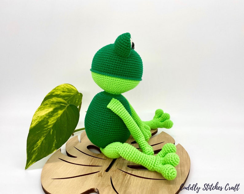 May include: A green crocheted frog sitting on a wooden leaf-shaped base. The frog has black eyes and a light green belly.