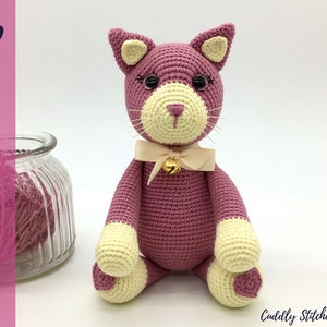 Peut inclure: Un chat en crochet rose avec des accents jaunes, portant un nœud blanc avec une clochette dorée. Le chat est assis sur un fond blanc.