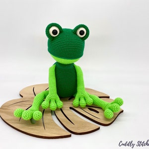May include: A green crocheted frog sitting on a wooden leaf-shaped base. The frog has large black eyes and a light green belly.
