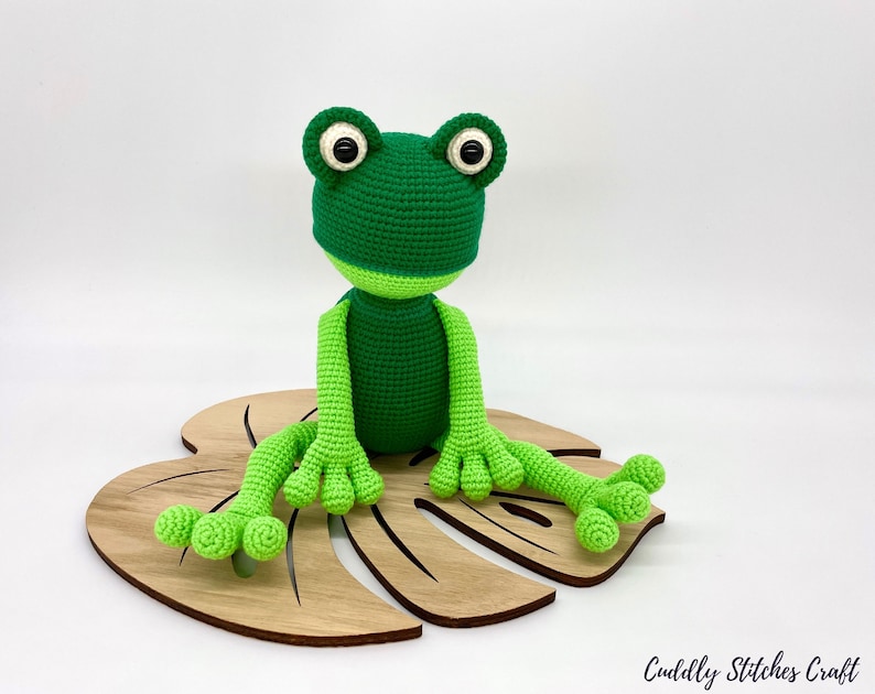 May include: A green crocheted frog sitting on a wooden leaf-shaped base. The frog has large black eyes and a light green belly.