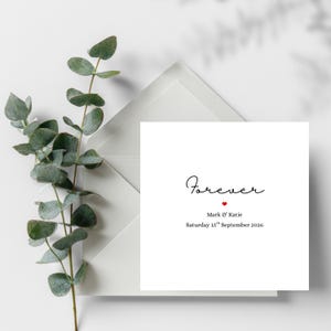 Könnte beinhalten: Eine weiße Hochzeitseinladungskarte mit dem Wort "Forever" in eleganter Schrift, einem kleinen roten Herz und den Namen "Mark & Katie" mit dem Datum "Samstag, 15. September 2026". Die Karte befindet sich in einem weißen Umschlag, mit Eukalyptusblättern im Hintergrund.