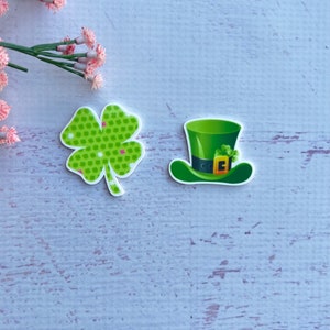 May include: Two green St. Patrick's Day themed resin embellishments. One is a four-leaf clover with a green and white polka dot pattern. The other is a green leprechaun hat with a gold buckle.