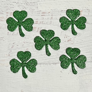 以下が含まれることがあります： 白い木製の背景に、緑色のグリッターでできたクローバーの切り抜きが5つある。