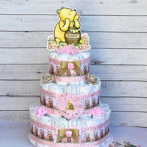 May include: A three-tiered diaper cake decorated with Winnie the Pooh and honey pot themed decorations. The cake is decorated with pink and gold ribbons, flowers, and a Winnie the Pooh topper.