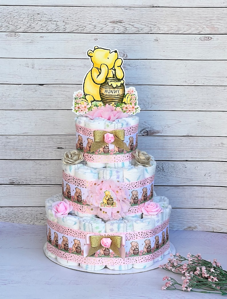 May include: A three-tiered diaper cake decorated with Winnie the Pooh and honey pot themed decorations. The cake is decorated with pink and gold ribbons, flowers, and a Winnie the Pooh topper.