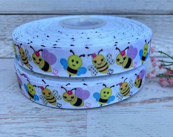 May include: White ribbon with a repeating pattern of cartoon bees with pink hearts and black polka dots. The ribbon is rolled up and ready to be used for crafts or decorations.