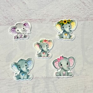 Könnte beinhalten: Fünf graue Elefantenfiguren mit verschiedenen farbigen Akzenten. Jeder Elefant sitzt mit seinem Rüssel eingerollt. Die Elefanten haben verschiedene Blumenkronen und Tupfenakzente.
