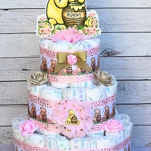 May include: A three-tiered diaper cake decorated with Winnie the Pooh and honey pot themed decorations. The cake is decorated with pink and gold ribbons, flowers, and Winnie the Pooh characters. The top tier features a Winnie the Pooh cutout holding a honey pot with the word "Hunny" on it.