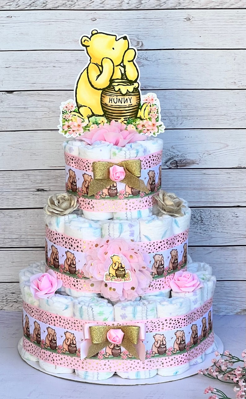 May include: A three-tiered diaper cake decorated with Winnie the Pooh and honey pot themed decorations. The cake is decorated with pink and gold ribbons, flowers, and Winnie the Pooh characters. The top tier features a Winnie the Pooh cutout holding a honey pot with the word "Hunny" on it.