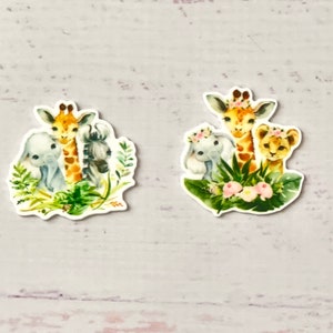 Könnte beinhalten: Zwei Sätze Tieraufkleber mit einer Giraffe, einem Elefanten und einem Löwen. Die Aufkleber sind von grünen Blättern und rosa Blumen umgeben.