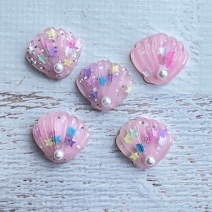 Pode incluir: Cinco pingentes de resina em forma de concha rosa com um detalhe de pérola. Cada pingente é cheio de glitter e estrelas coloridas.