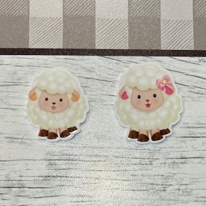 May include: Two white fluffy sheep with brown hooves and black eyes. One sheep has a pink flower on its head.