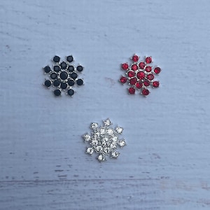 May include: Three snowflake-shaped embellishments with rhinestones. The embellishments are silver with black, red, and clear rhinestones.