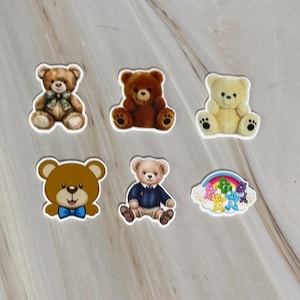 May include: A collection of six stickers featuring various teddy bears. One bear wears a green bow, another a blue bow, one is dressed in a sweater and shorts, and one sticker depicts a group of small colourful bears beneath a rainbow.