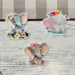 Könnte beinhalten: Drei Aquarell-Elefantenillustrationen mit floralen Akzenten. Jeder Elefant sitzt und hat eine rosa Nase und einen grauen Körper mit weißen Flecken. Die Elefanten sind von Blumen und Grün umgeben.