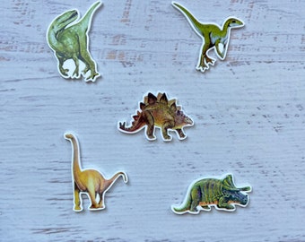 Może przedstawiać: Sześć kolorowych naklejek z dinozaurami na białym tle. Dinozaury to zielony welociraptor, zielony stegozaur, brązowy stegozaur, żółty brachiozaur, zielony triceratops i zielony welociraptor.