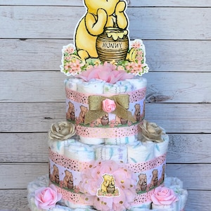 May include: A three-tiered diaper cake decorated with pink and gold ribbons, flowers, and Winnie the Pooh characters. The top tier features a Winnie the Pooh cutout holding a honey pot with the word "Hunny" on it.