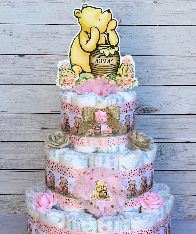 May include: A three-tiered diaper cake decorated with pink and gold ribbons, flowers, and Winnie the Pooh characters. The top tier features a Winnie the Pooh cutout holding a honey pot with the word "Hunny" on it.