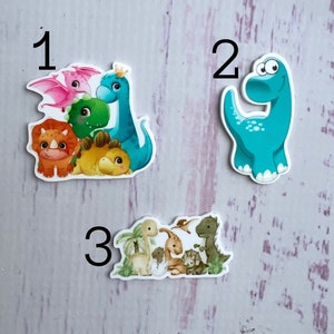 Może przedstawiać: Trzy zestawy kolorowych naklejek z dinozaurami. Pierwszy zestaw zawiera grupę dinozaurów, w tym różowego dinozaura z koroną. Drugi zestaw zawiera jednego niebieskiego dinozaura. Trzeci zestaw zawiera grupę dinozaurów z palmą.