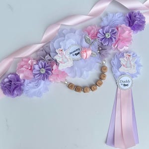 May include: A maternity sash and matching badge set featuring light purple and pink fabric flowers, satin ribbons, and a small pink rose. The sash includes wooden letter beads spelling ANELLIE and a circular tag reading Mommy to be with a dragon graphic.