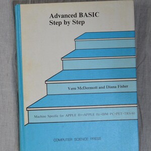 May include: A softcover book titled Advanced BASIC Step by Step by Vern McDermott and Diana Fisher. The cover features a blue staircase graphic on a white background, with text indicating it is for Apple II+, Apple IIe, IBM PC, PET, and TRS-80.