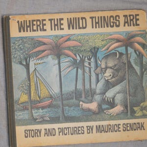 May include: A hardcover children's book titled Where the Wild Things Are by Maurice Sendak. The cover features a large, furry, horned creature sitting near a sailboat on a river, surrounded by stylized trees, all rendered in muted earth tones.