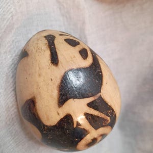 May include: An oval-shaped object with a light tan, matte surface featuring dark brown, burnt-in abstract markings. The design consists of curved, organic shapes that wrap around the rounded form, creating a high-contrast pattern across the exterior.