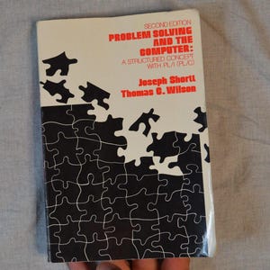 May include: A softcover book titled Problem Solving and the Computer: A Structured Concept with PL/I (PL/C) by Joseph Shortt and Thomas C. Wilson. The white cover features a black puzzle piece graphic pattern and red text for the title.