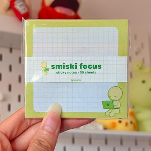 May include: A square pad of light green sticky notes with a white grid pattern. The paper band features the text smiski focus sticky notes 50 sheets lylanite and a small illustration of a green character using a laptop computer.