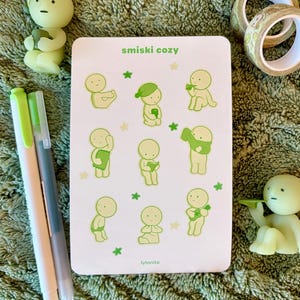 May include: A white rectangular card with the text "smiski cozy" and illustrations of small, green figures in various poses. Two green pens, two small figurines, and decorative tape rolls are also visible.