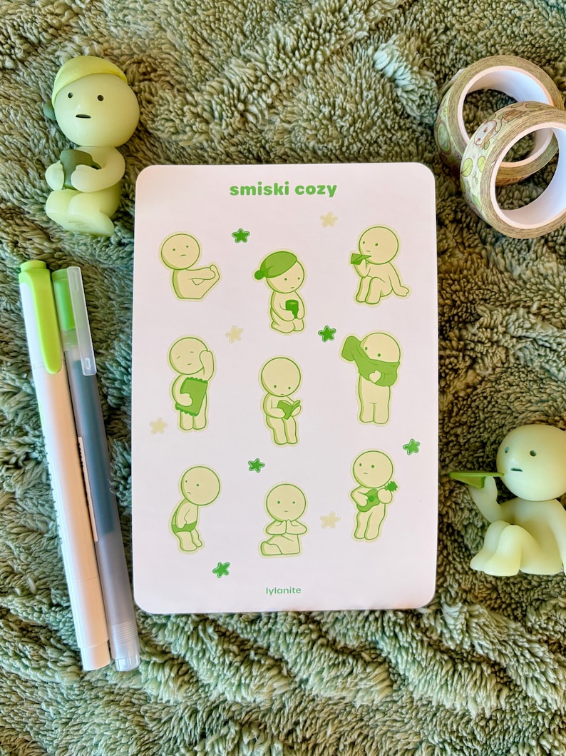 May include: A white rectangular card with the text "smiski cozy" and illustrations of small, green figures in various poses. Two green pens, two small figurines, and decorative tape rolls are also visible.