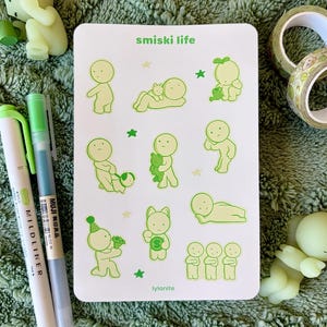 May include: A white sticker sheet with green illustrations of small figures in various poses, with the text "smiski life" and "lylanite". Two green and white pens and two small green figurines are also visible.