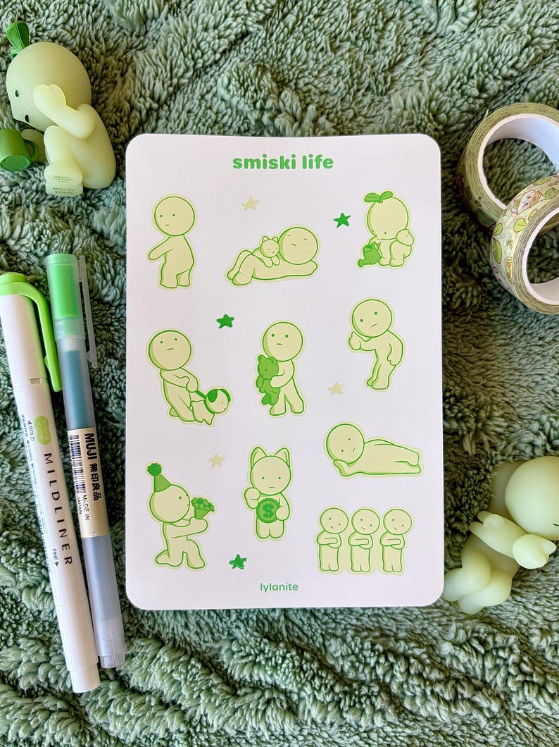 May include: A white sticker sheet with green illustrations of small figures in various poses, with the text "smiski life" and "lylanite". Two green and white pens and two small green figurines are also visible.