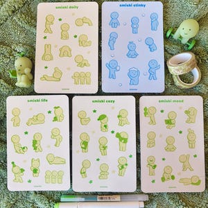 May include: A collection of Smiski figurines and sticker sheets. The sticker sheets are labeled "smiski daily", "smiski stinky", "smiski life", "smiski cozy", and "smiski mood". The figurines and stickers are in shades of green and blue.