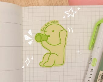 May include: A light green sticker featuring a cartoon character sitting and drinking from a green bottle, with the text "You got this!" arched above its head. The sticker is placed on a white grid-patterned notebook page next to a pen.