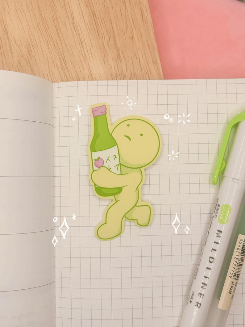 May include: A die-cut sticker featuring a light green, rounded character holding a green bottle with a pink cap and a peach illustration. The sticker is placed on a grid-patterned notebook page next to a light green Mildliner marker.