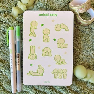 May include: A collection of stationery items on a green surface. Includes a white card with green illustrations and text that reads "smiski daily" and "lylanite". Also features green pens, washi tape, and small figurines.