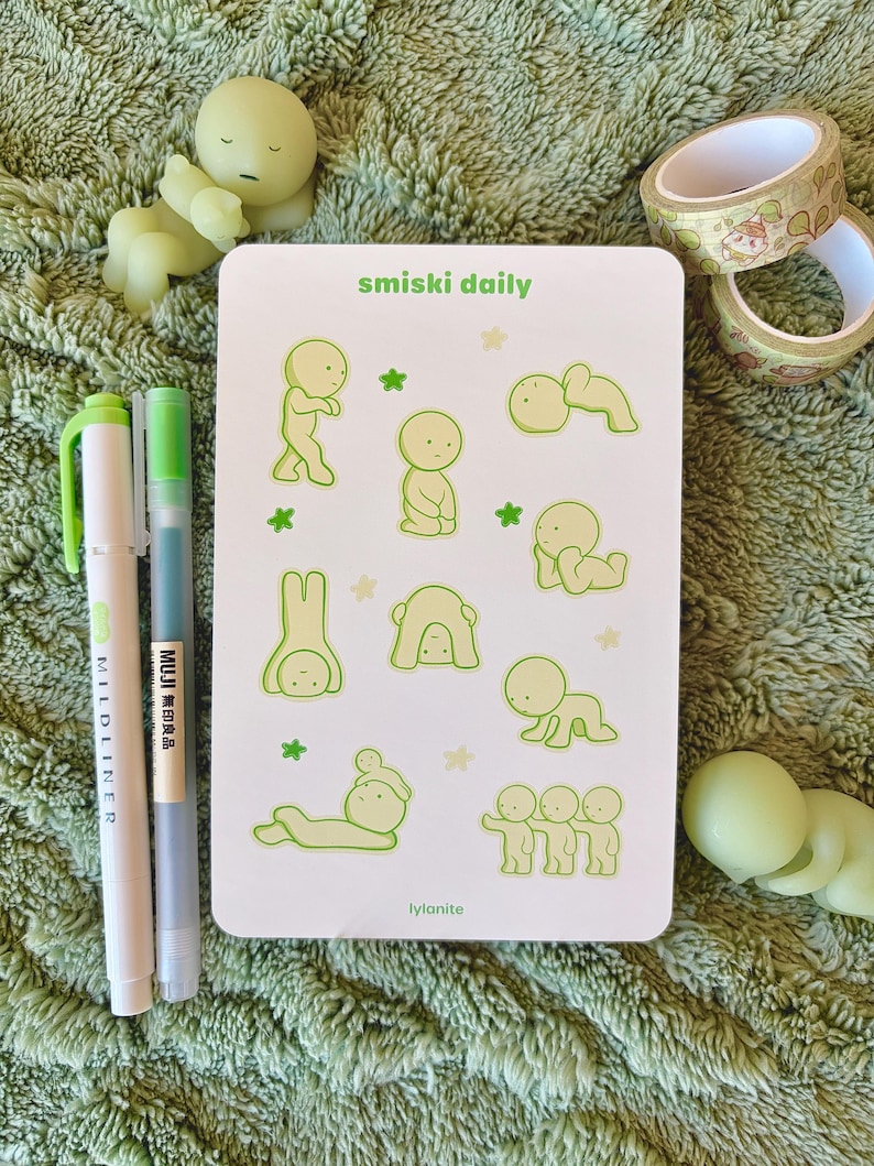 May include: A collection of stationery items on a green surface. Includes a white card with green illustrations and text that reads "smiski daily" and "lylanite". Also features green pens, washi tape, and small figurines.