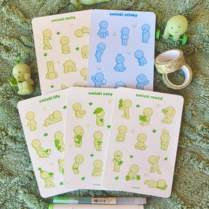 May include: A collection of Smiski figurines and sticker sheets. The sticker sheets feature various poses of small, cartoon-like figures in green and blue. The figurines are in various poses, and there are two pens and decorative tape rolls.