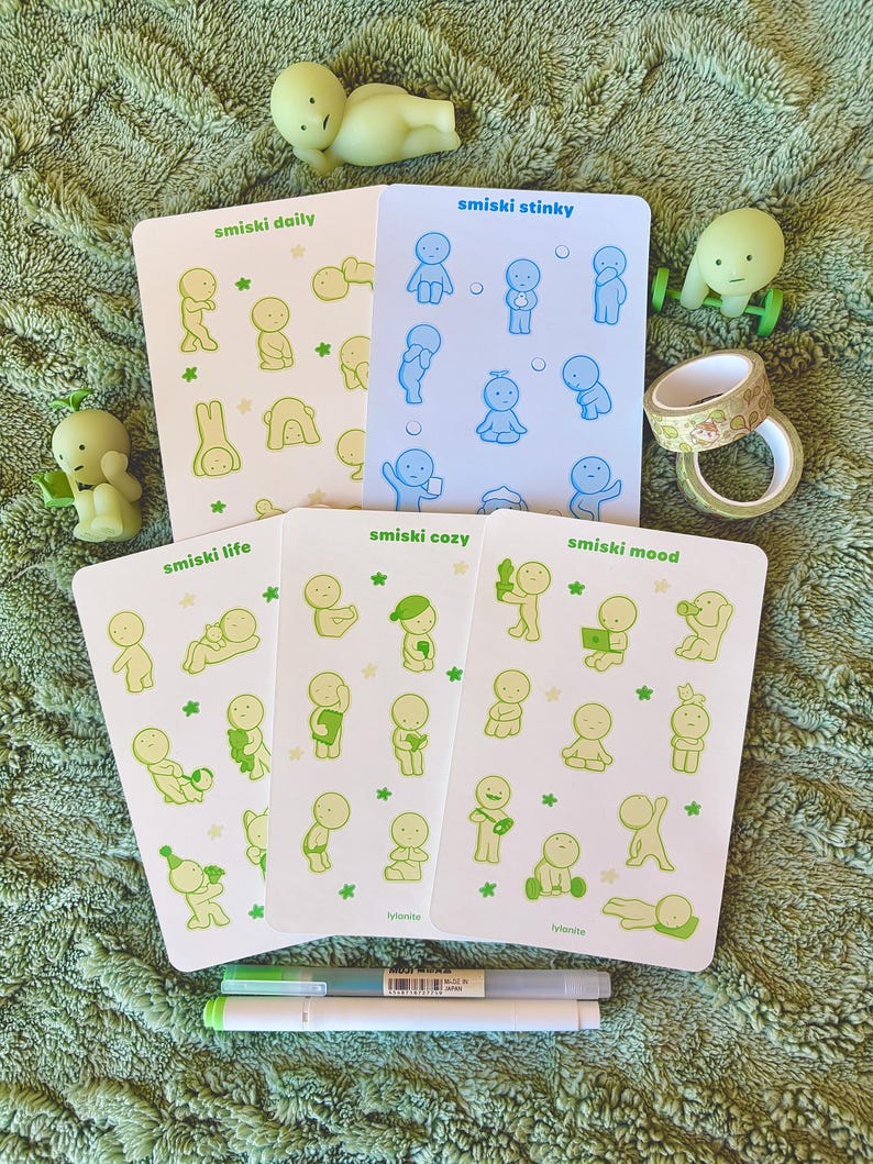 May include: A collection of Smiski figurines and sticker sheets. The sticker sheets feature various poses of small, cartoon-like figures in green and blue. The figurines are in various poses, and there are two pens and decorative tape rolls.