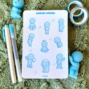 May include: A collection of stationery items on a green textured surface. The items include a white card with blue cartoon figures and the text "smiski stinky", two blue pens, two blue figurines, and two rolls of decorative tape.