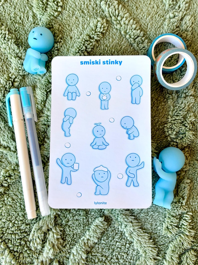 May include: A collection of stationery items on a green textured surface. The items include a white card with blue cartoon figures and the text "smiski stinky", two blue pens, two blue figurines, and two rolls of decorative tape.